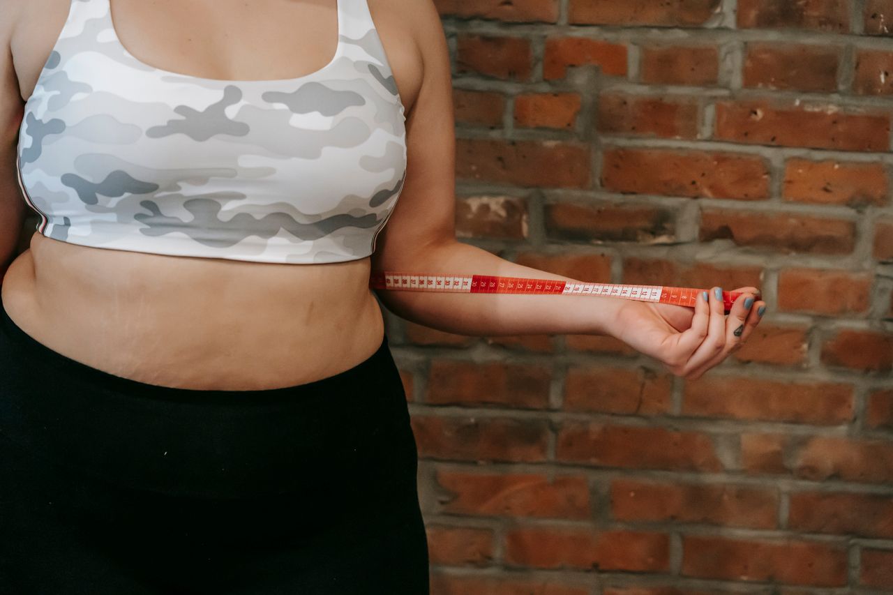 About Crop anonymous plump female using measuring tape for measuring belly near brick wall