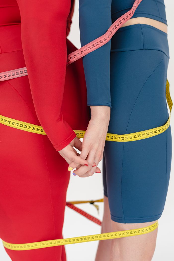 Side view of unrecognizable plus size female with slender woman with measuring tapes standing back to back on white background